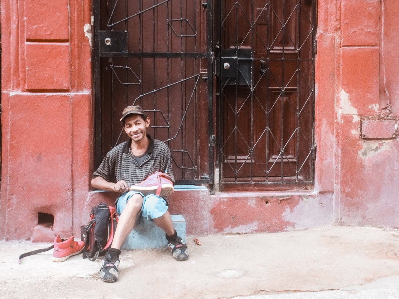 Smiling man sits in front of a red urban door, holding a shoe, capturing a moment of street life.