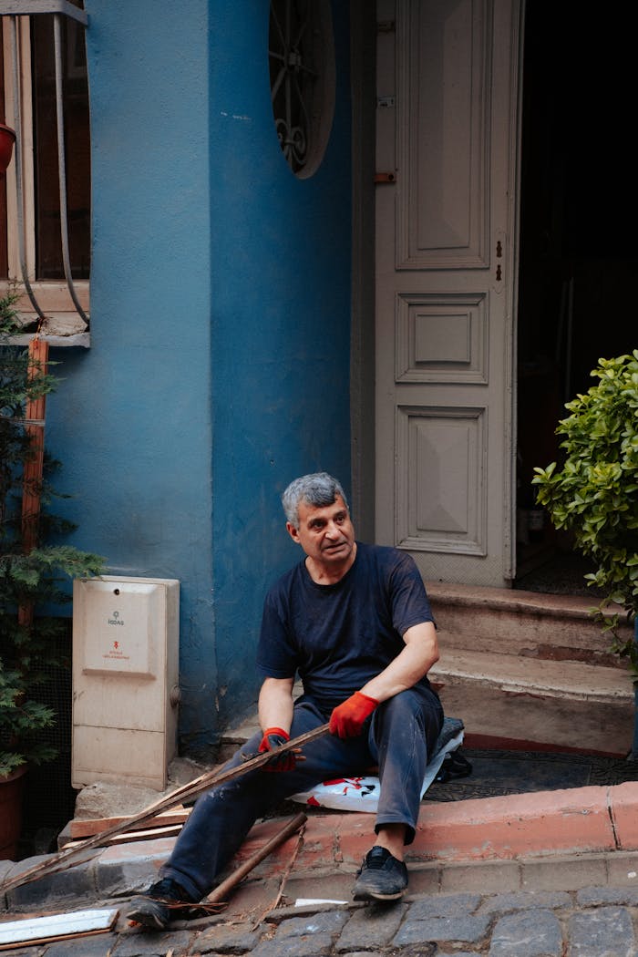 Elderly man in casual attire sitting outdoors near a blue building entrance, holding tools.